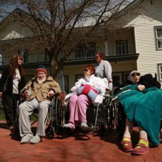 A group of elderly individuals in wheelchairs with caregivers outside a building.