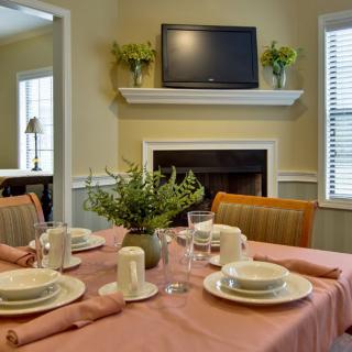 A cozy dining area with a neatly set table featuring a pink tablecloth and white dinnerware, positioned near a fireplace with a television above it.