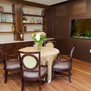 A cozy dining room with a round table and floral centerpiece, surrounded by elegant chairs. The room includes wooden flooring, built-in shelves with decorative items, and an aquarium embedded in the wall.