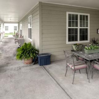 A covered outdoor patio area with a table, chairs, and potted plants along a concrete walkway.