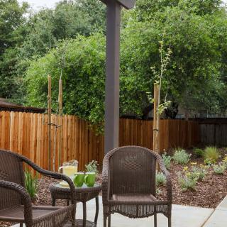 A cozy patio area with wicker chairs and a small table, surrounded by a garden and wooden fence.
