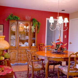 A cozy dining room with a wooden table set, surrounded by classic chairs and a chandelier hanging above. The room features red walls and a display cabinet filled with decorative items.