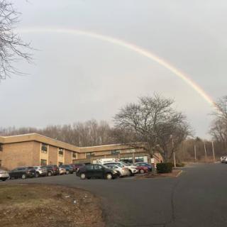 A beautiful rainbow arcs over a parking lot beside a building on a cloudy day.
