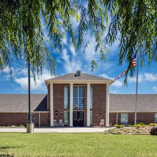 A brick building with a flag in front, surrounded by a green lawn and trees, under a blue sky.