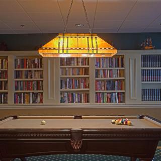 A billiards room with a pool table under a warm light fixture, surrounded by bookshelves filled with books.