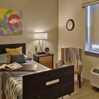 A cozy bedroom with modern decor, featuring a bed, side table with a lamp, colorful wall art, and an upholstered chair by the window.