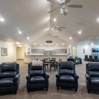 A spacious modern living room with four black recliners, a dining area in the background, and ceiling fans overhead.