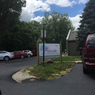 Entrance to a facility with a parking area surrounded by trees under a partly cloudy sky.