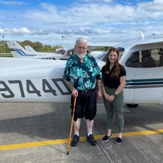 Two people standing in front of a small airplane on a tarmac.