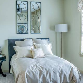 A cozy bedroom featuring a neatly made bed with multiple pillows, decorative mirrors above the headboard, and a floor lamp.