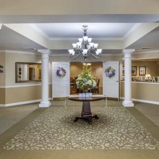A well-decorated lobby featuring a central table with floral arrangements, illuminated by a chandelier and flanked by columns.