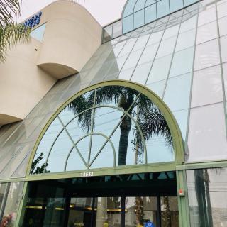 A modern shopping mall entrance with a glass facade and palm trees reflected in the windows.
