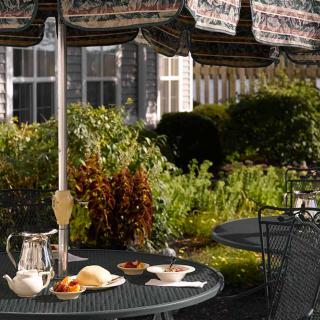 A peaceful outdoor patio setting with a table set for breakfast, featuring an umbrella and surrounded by a garden.