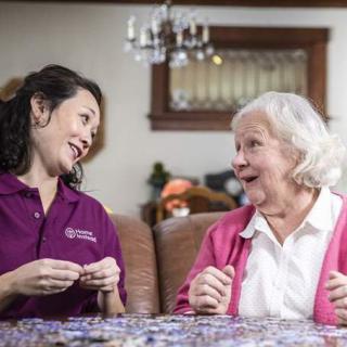 A caregiver and an elderly woman share a joyful conversation in a cozy living room setting.