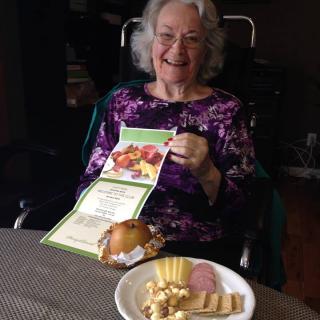 An elderly woman in a wheelchair smiles while holding a card, with a plate of food in front of her.