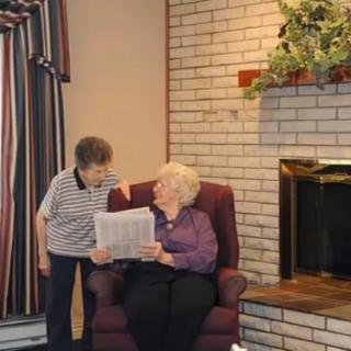 Two elderly individuals share a conversation in a cozy living room, one seated and holding a newspaper.