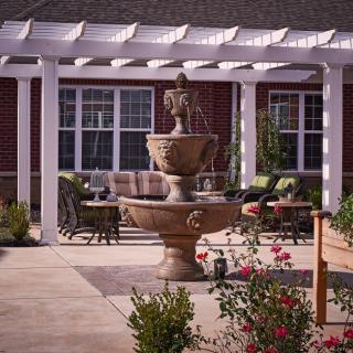 A serene outdoor garden patio with a central stone fountain, surrounded by seating and vibrant flowers under a pergola.