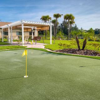 A well-maintained putting green with a flag, surrounded by landscaping and palm trees, next to a building.