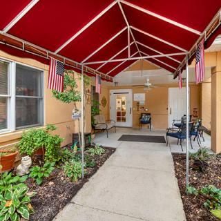 A covered entrance with a red awning, bordered by plants and flags, leads to a doorway.