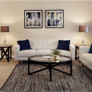 A cozy living room with two white sofas, a round coffee table, and stylish lamps on end tables. The walls feature modern artwork above the seating area.
