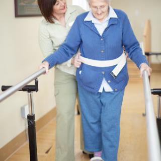 A caregiver assists an elderly woman as she practices walking using support rails.