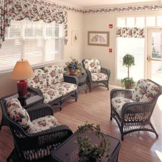 A cozy living room featuring wicker furniture with floral patterned cushions, warmly lit by a lampshade and enhanced by natural light from the windows.