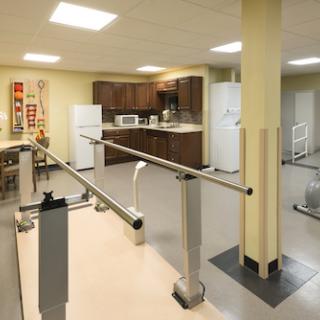 A bright and spacious rehabilitation therapy room featuring parallel bars and a kitchenette.
