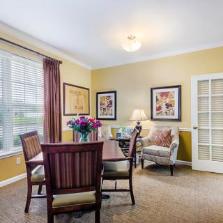 A cozy dining room with a wooden table and chairs, decorated with artwork and illuminated by a lamp near the window.