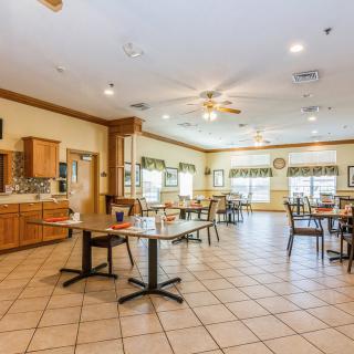 A spacious dining room with several tables and chairs, featuring a kitchen area and large windows allowing natural light.