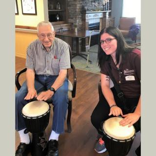 An elderly man and a young woman enjoying drumming together indoors.