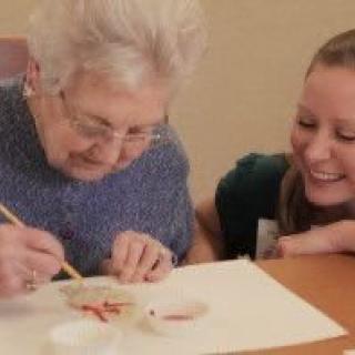 An elderly woman painting with a younger woman watching and smiling.