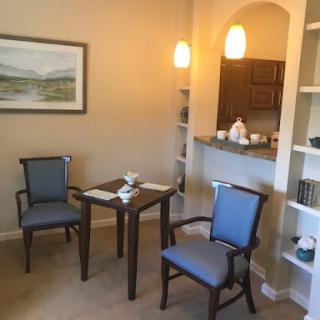 A cozy dining area featuring a small table and two chairs, with a well-lit kitchen in the background.