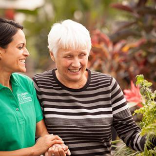 A caregiver and an elderly woman enjoy gardening together outdoors.