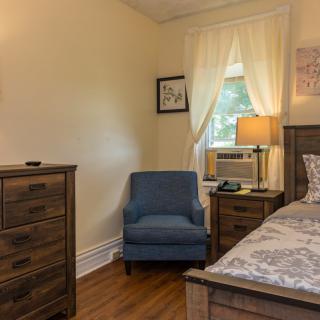A cozy bedroom with a blue armchair, wooden dresser, and bed near a window with white curtains.