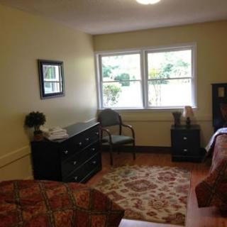A cozy bedroom with a window view, featuring a dresser, chair, and patterned carpet.