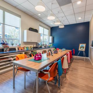 A modern art classroom with colorful chairs and backpacks, featuring large windows and a blue accent wall.