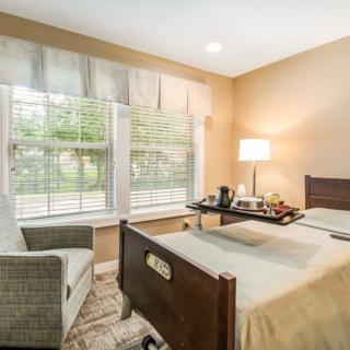 A cozy bedroom with a hospital bed, well-lit by a nearby lamp, featuring large windows and comfortable seating.