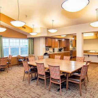 A well-lit dining area with a large table surrounded by chairs, adjacent to a kitchen.