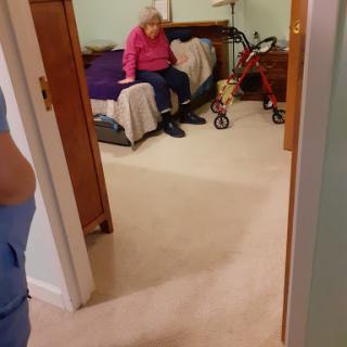 A cozy bedroom scene featuring an elderly person sitting on the bed while a caregiver is in the foreground.