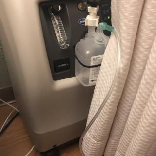 A portable oxygen concentrator with a connected humidifier bottle and tube, placed indoors.