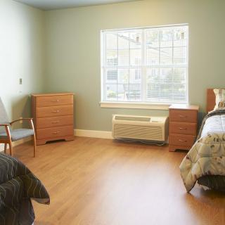A minimalist bedroom with a bed, two dressers, and a chair, featuring a large window and wooden floors.