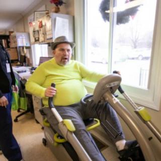 An elderly person is using fitness equipment indoors, wearing a hat and a bright green shirt.