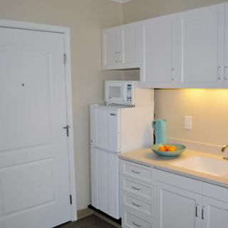 A compact kitchen area with white cabinets and appliances, including a microwave and fridge, with a countertop featuring a bowl of fruit and a ceramic vase.