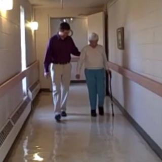 An elderly person is assisted by a caregiver while walking down a hallway.
