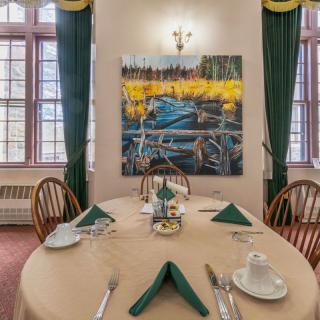 A cozy dining room with an elegantly set table, featuring a vibrant painting and large windows adorned with green curtains.