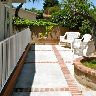 A sunny patio with white chairs, surrounded by a garden and brick pathways.