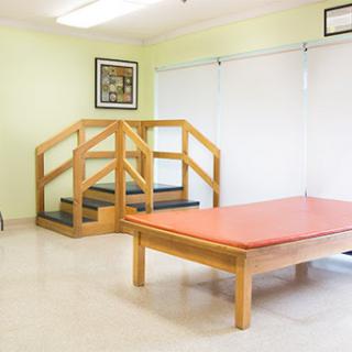 A spacious therapy room featuring a wooden table and stairs, designed for patient comfort.