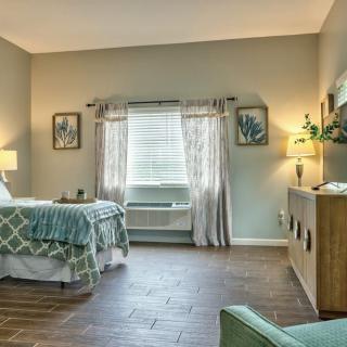 A cozy bedroom with a neatly made bed, framed pictures on the walls, a window with patterned curtains, and soft lighting from lamps.