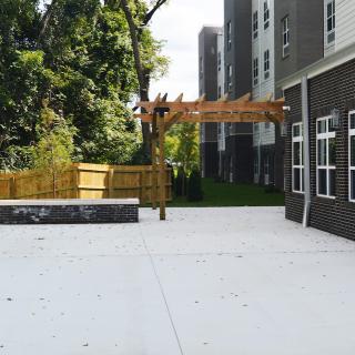 A modern outdoor patio area adjacent to a building, featuring a wooden pergola and surrounded by trees and a fence.