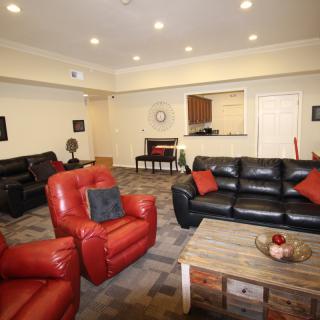 A cozy living room featuring red chairs and black sofas arranged around a wooden coffee table, with warm lighting and a carpeted floor.